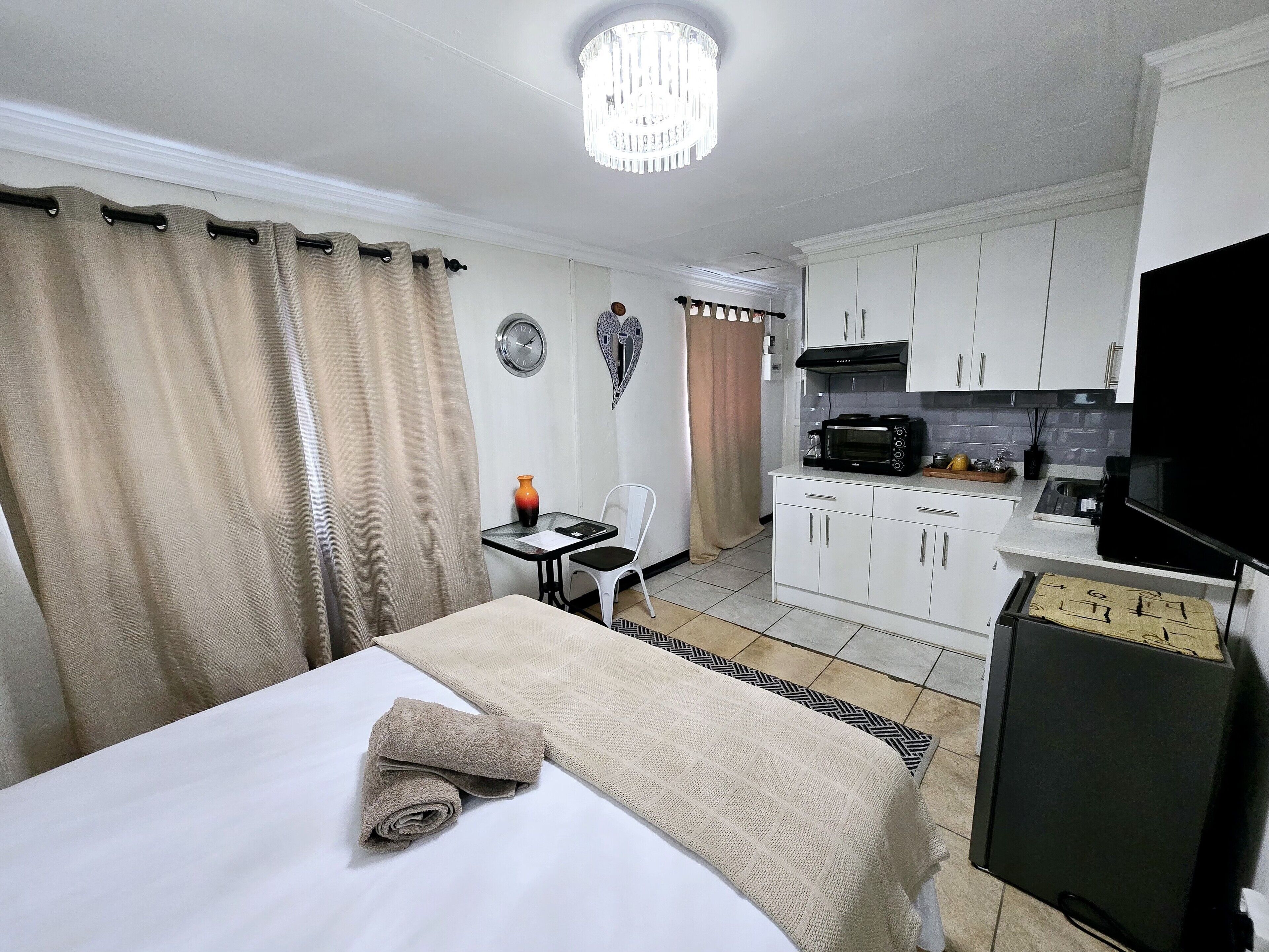 A hotel room with a bed, a small table with a chair, a kitchenette, and a TV.