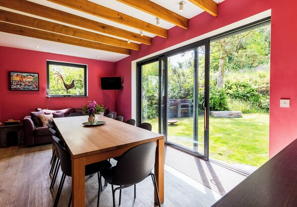 Un espace repas avec une table et des chaises en bois, un mur rouge, une grande fenêtre donnant sur la verdure et une œuvre d’art accrochée au mur.