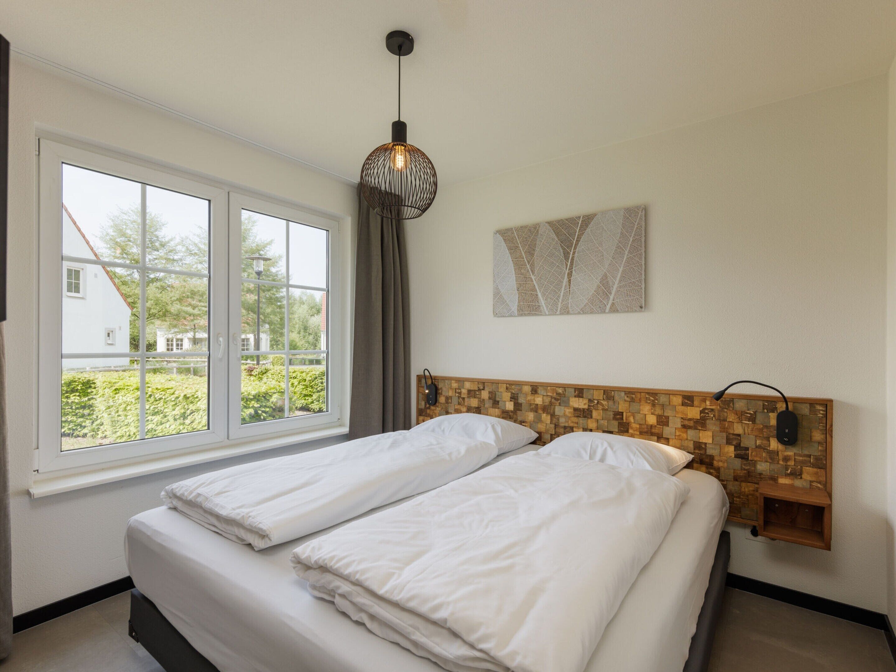 A bedroom with a large window, a bed with white linens, a hanging light fixture, and a wall-mounted speaker.