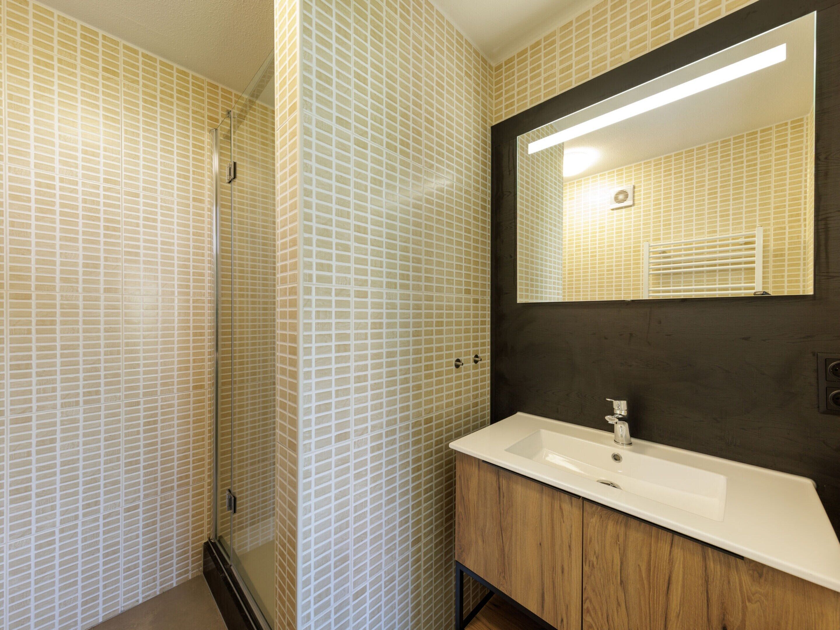A bathroom with a tiled wall, a wooden vanity, a white sink, and a mirror.