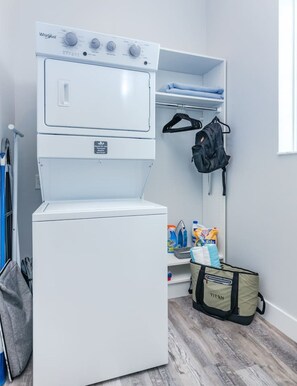 A compact laundry room with a Whirlpool washer and dryer, a drying rack, and storage shelves with cleaning supplies.