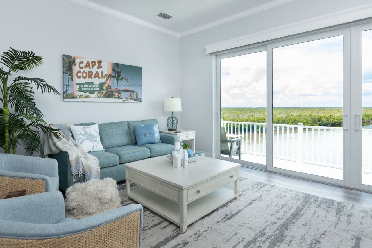 A modern living room with a large window, a sofa, a coffee table, and a painting of a coastal scene.