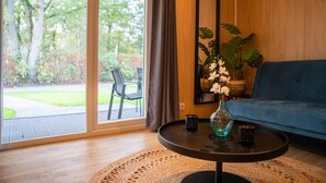 Una habitación con un gran ventanal corredero que da a un jardín, un sofá azul, una mesa redonda con un jarrón y dos tazas, y un espejo grande en la pared.