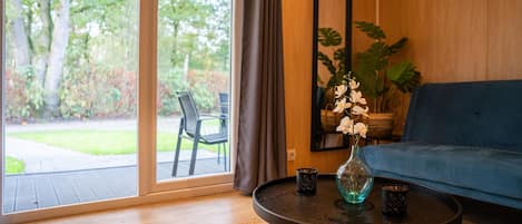 A room with a large sliding glass door leading to a garden, a blue sofa, a round table with a vase and two cups, and a large mirror on the wall.