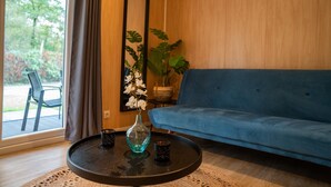 Una sala de estar con un sofá azul, una mesa de centro redonda con un jarrón y dos velas, un espejo grande y una planta en maceta.