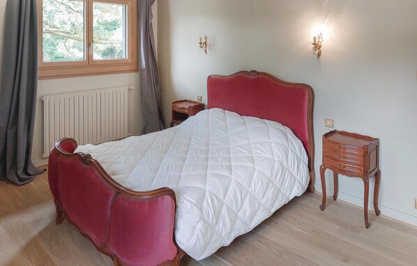 Un dormitorio con una cama tapizada en rojo, una mesita de noche de madera, una lámpara fijada a la pared y una ventana con cortinas.