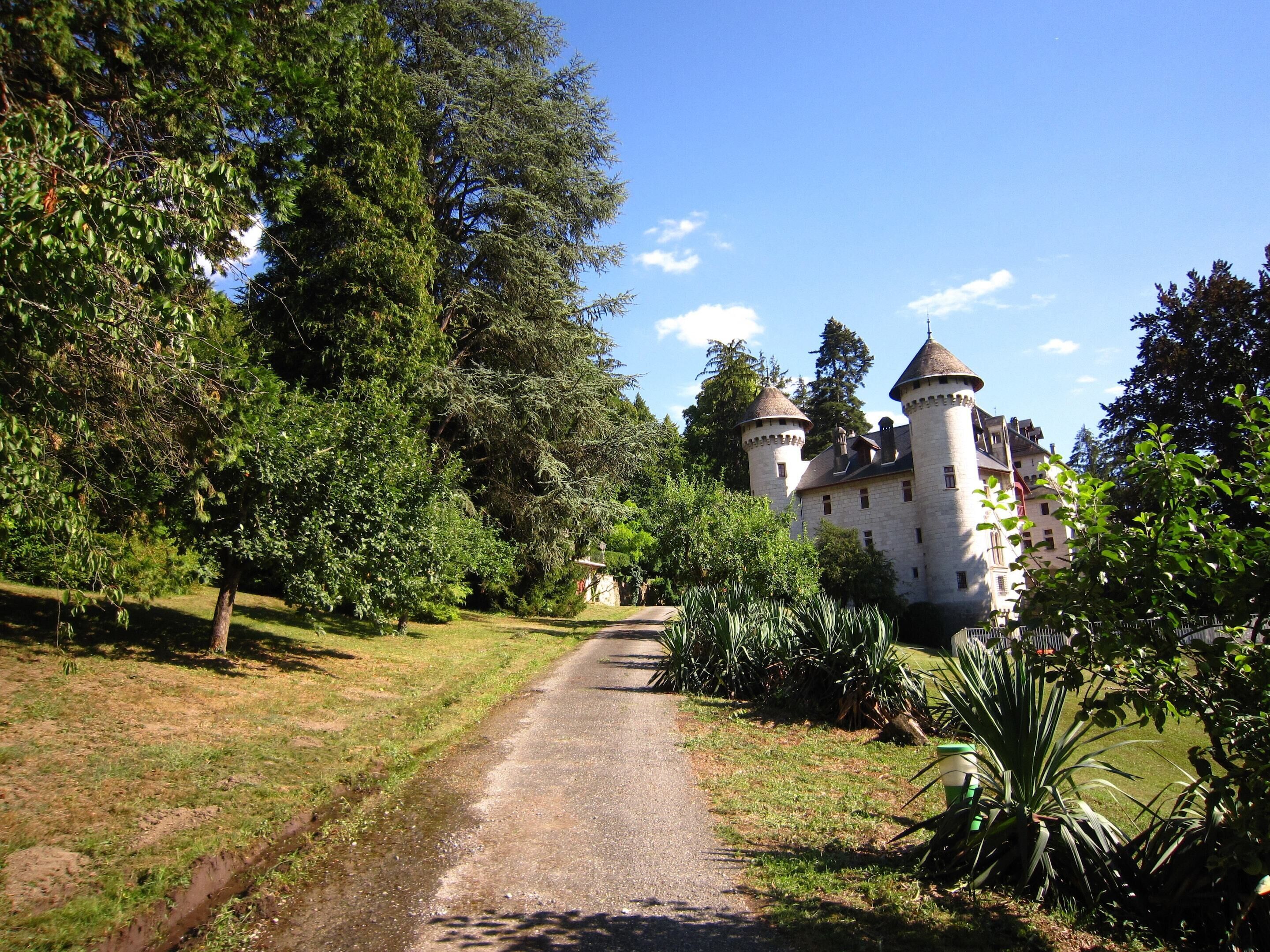 Un château doté de deux tours, entouré d’une végétation luxuriante et accessible par un chemin de gravier.