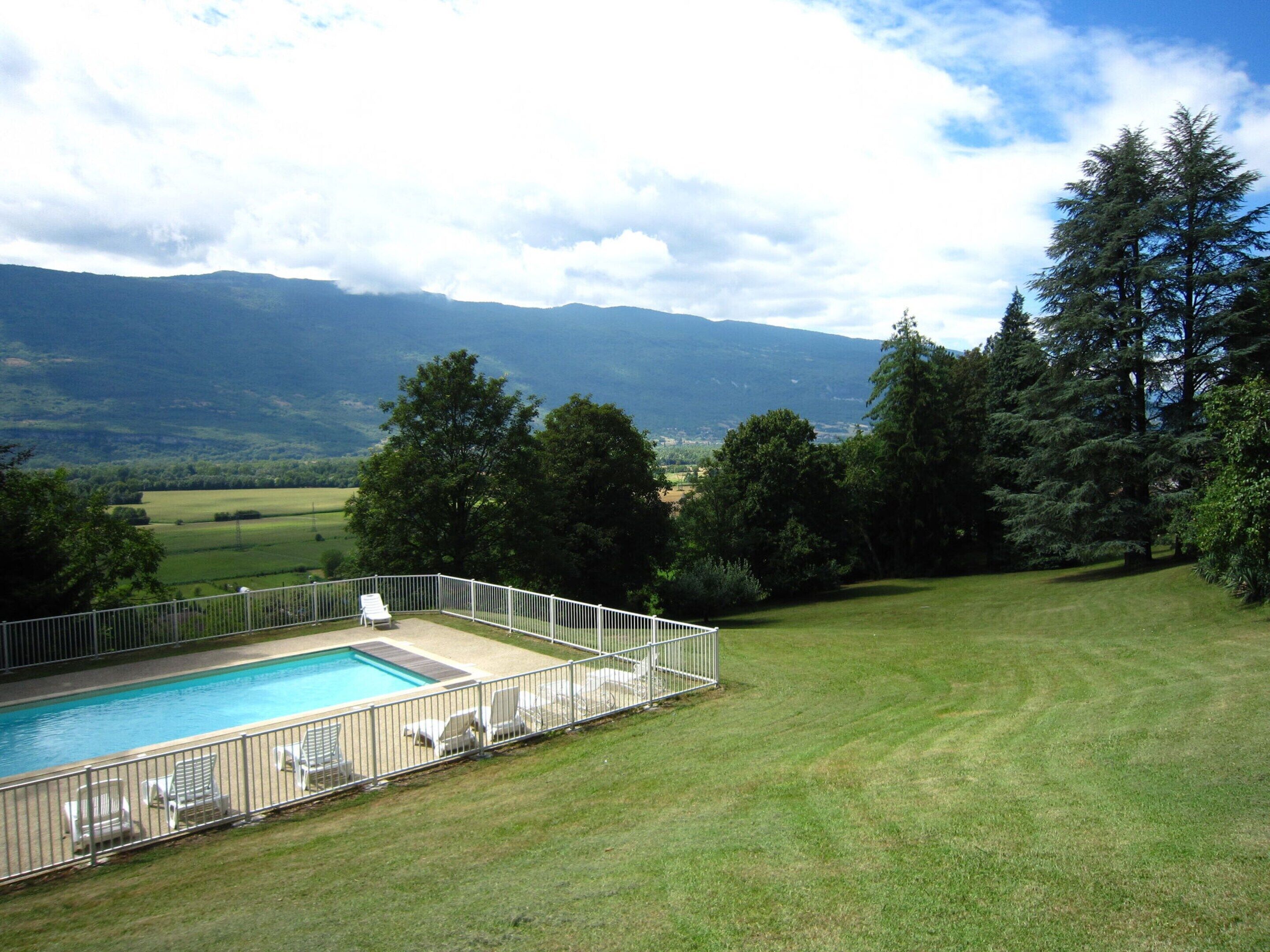 A swimming pool with lounge chairs, surrounded by a white fence, set against a backdrop of mountains and trees.