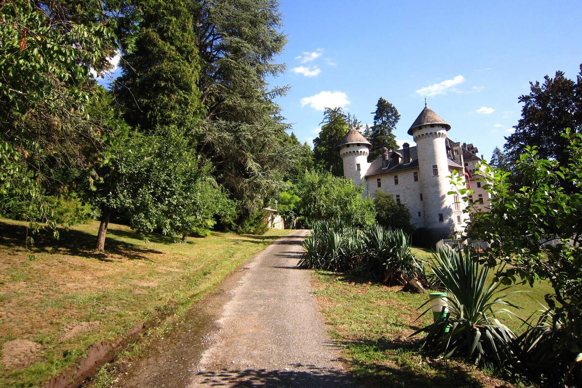 A castle with two towers, surrounded by lush greenery and a gravel path leading to it.