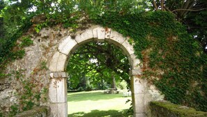 Un arco de piedra con musgo y hiedra, que conduce a un área verde con un banco y árboles.