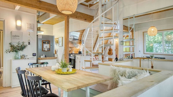 Interior de una cabaña con una mesa de comedor de madera, sillas, un banco, una escalera de caracol y una zona de cocina.