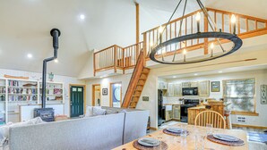 A spacious living room with a high ceiling, a wooden staircase, a dining area with a wooden table set for four, and a kitchen in the background. - Port Orchard Lakefront Cabin w/ Fire Pit & Grill! (Port Orchard)