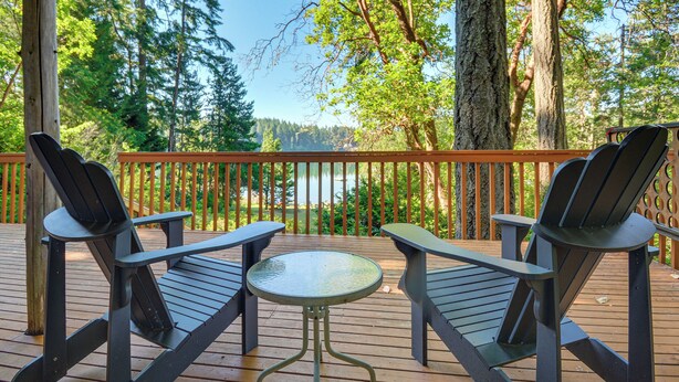 A wooden deck with two Adirondack chairs and a small table, overlooking a forested area and a body of water. - Port Orchard Lakefront Cabin w/ Fire Pit & Grill! (Port Orchard)