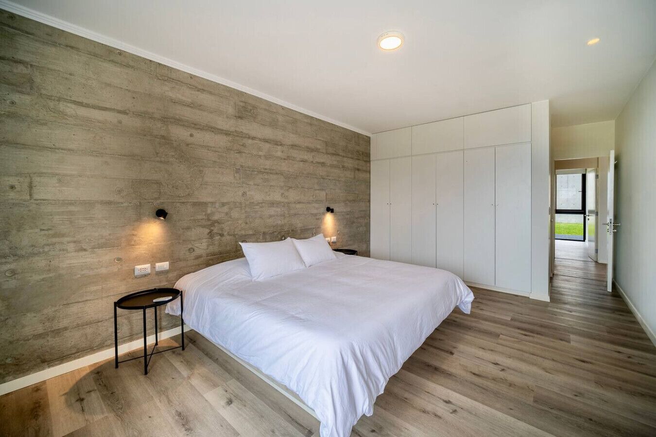 A bedroom with a large bed, wooden flooring, a concrete accent wall, and a wardrobe.