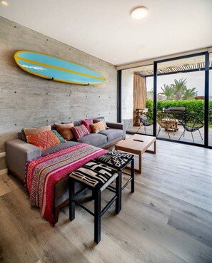 A modern living room with a grey sofa, colorful cushions, a wooden coffee table, and a surfboard on the wall. - Spacious apartment w/ Terrace 5min from Kite Point, the Bay and National Reserve (Paracas)