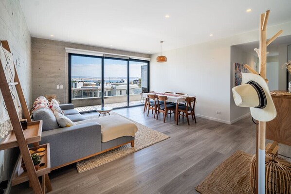 A modern living room with a sofa, dining table, and chairs. There is a large window with a view of buildings and the sky. - Beautifully Decorated Apt with Ocean View, Balcony & Kamado next to the Bay (Paracas)