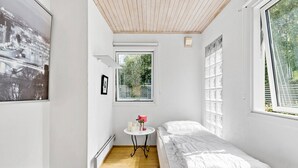 A small, bright room with a single bed, a small table with a lamp, and a window with blinds.