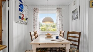 A dining room with a wooden table, chairs, a bowl of fruit, a blue cup, and a window with curtains.