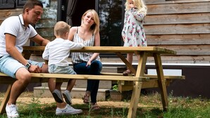 Une famille de quatre personnes est assise à une table en bois à l’extérieur, devant une cabane en bois sur laquelle est affiché un panneau indiquant « WOODLODE CL ».