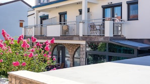 Un immeuble à plusieurs étages doté de balcons, d’arcades en pierre et d’un jardin où poussent des fleurs roses.
