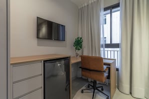 Habitación compacta con televisión de pantalla plana en la pared, un refrigerador pequeño, un escritorio de madera, una silla de oficina marrón y una planta en maceta.