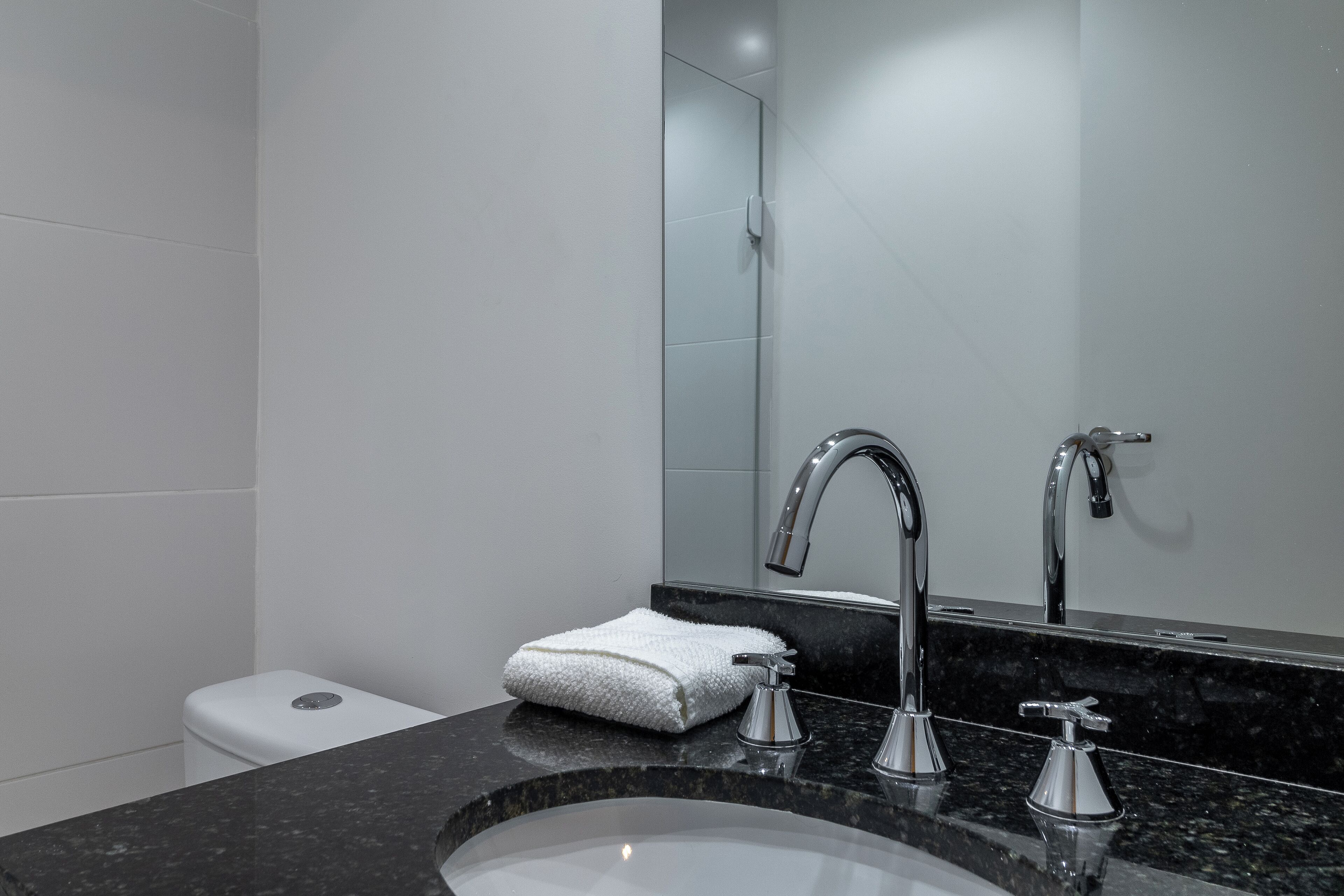 Un baño moderno con lavamanos de granito negro, dos grifos cromados, inodoro blanco y una toalla blanca sobre el mostrador.