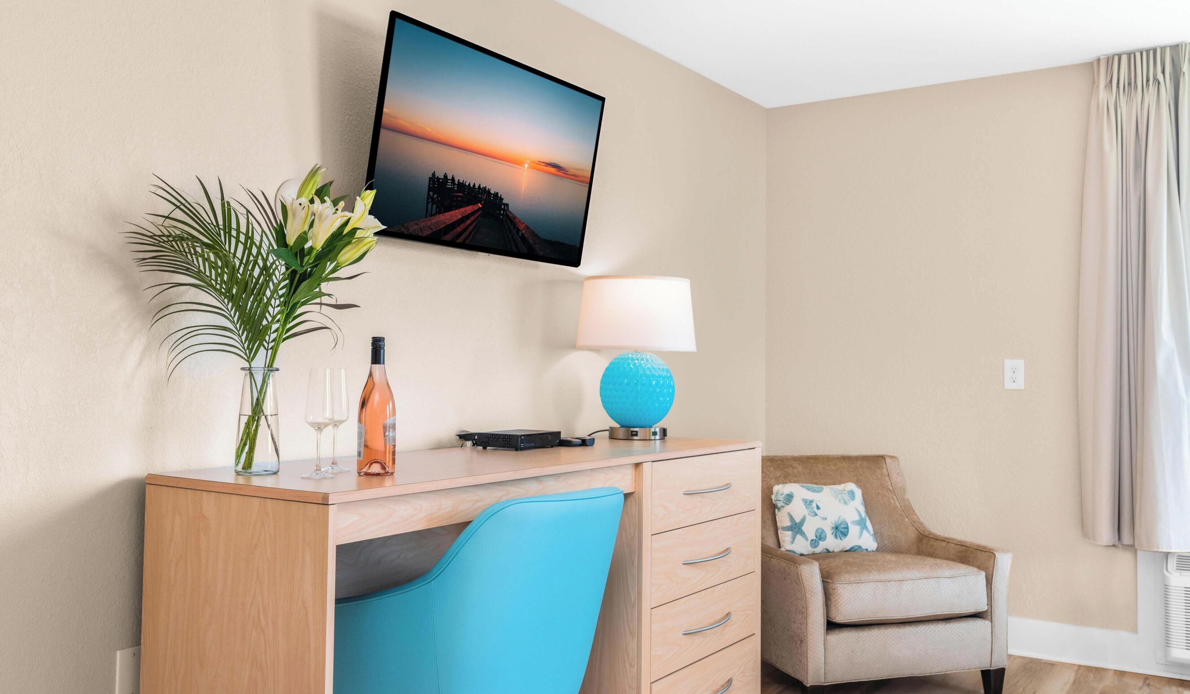Habitación de hotel con escritorio de madera, televisor montado en la pared, lámpara, silla y ventana con cortinas.