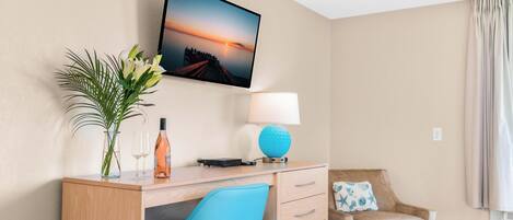 A hotel room with a wooden desk, a television mounted on the wall, a lamp, a chair, and a window with curtains.