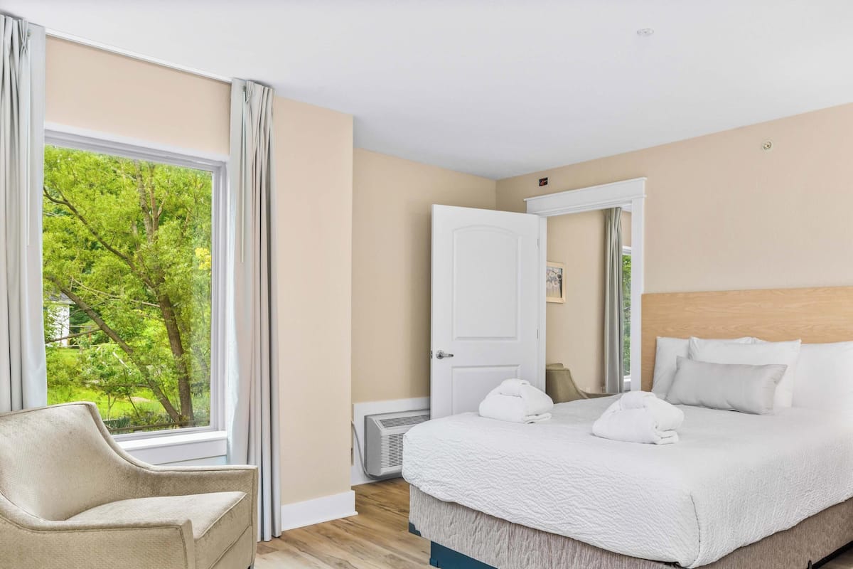 A hotel room with a bed, a chair, a bedside table, and a window with a view of trees.