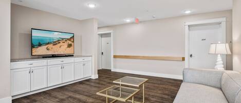 A modern living room with a flat-screen TV, a glass-top coffee table, a grey sofa, and a wooden bench.