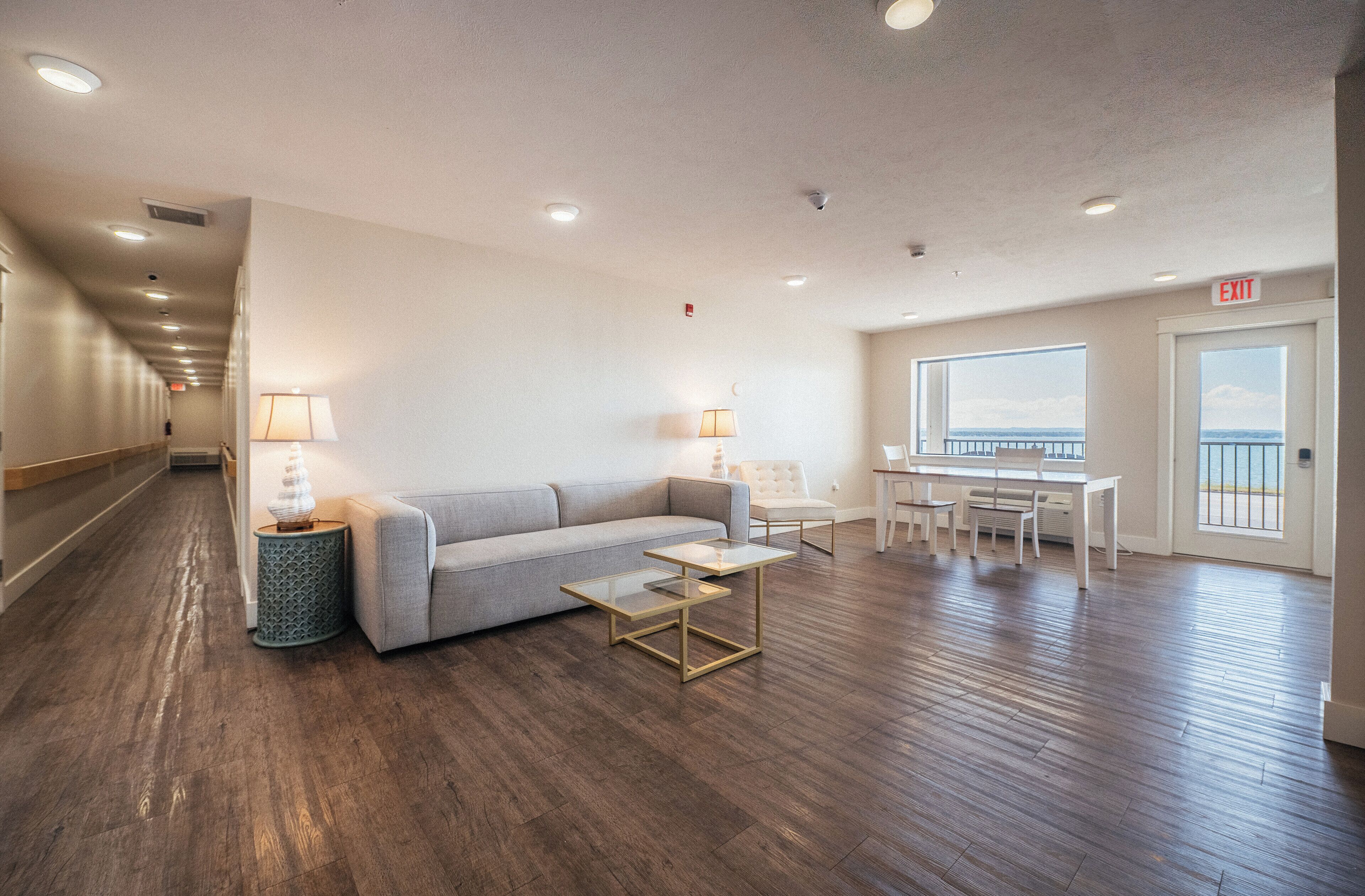 A hotel room with a sofa, two tables, a lamp, and a view of the sea through a window.