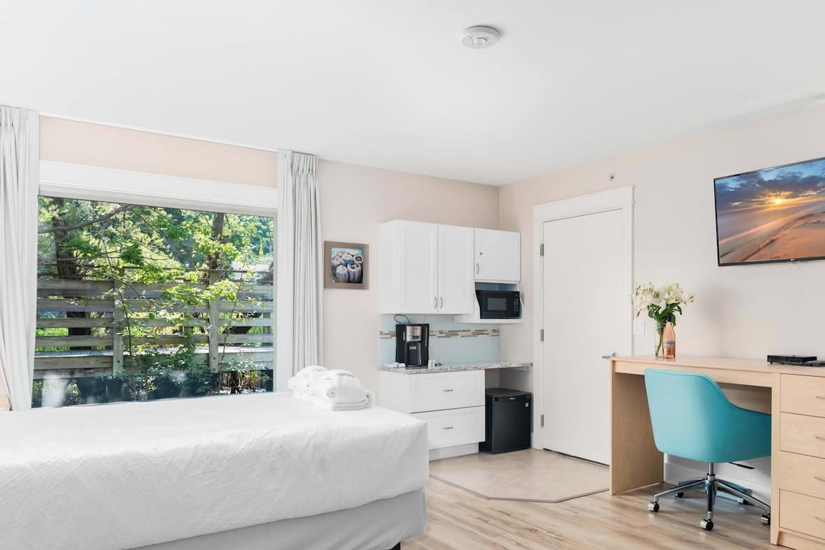 A bedroom with a bed, a desk, a chair, a small kitchenette, and a view of greenery outside.