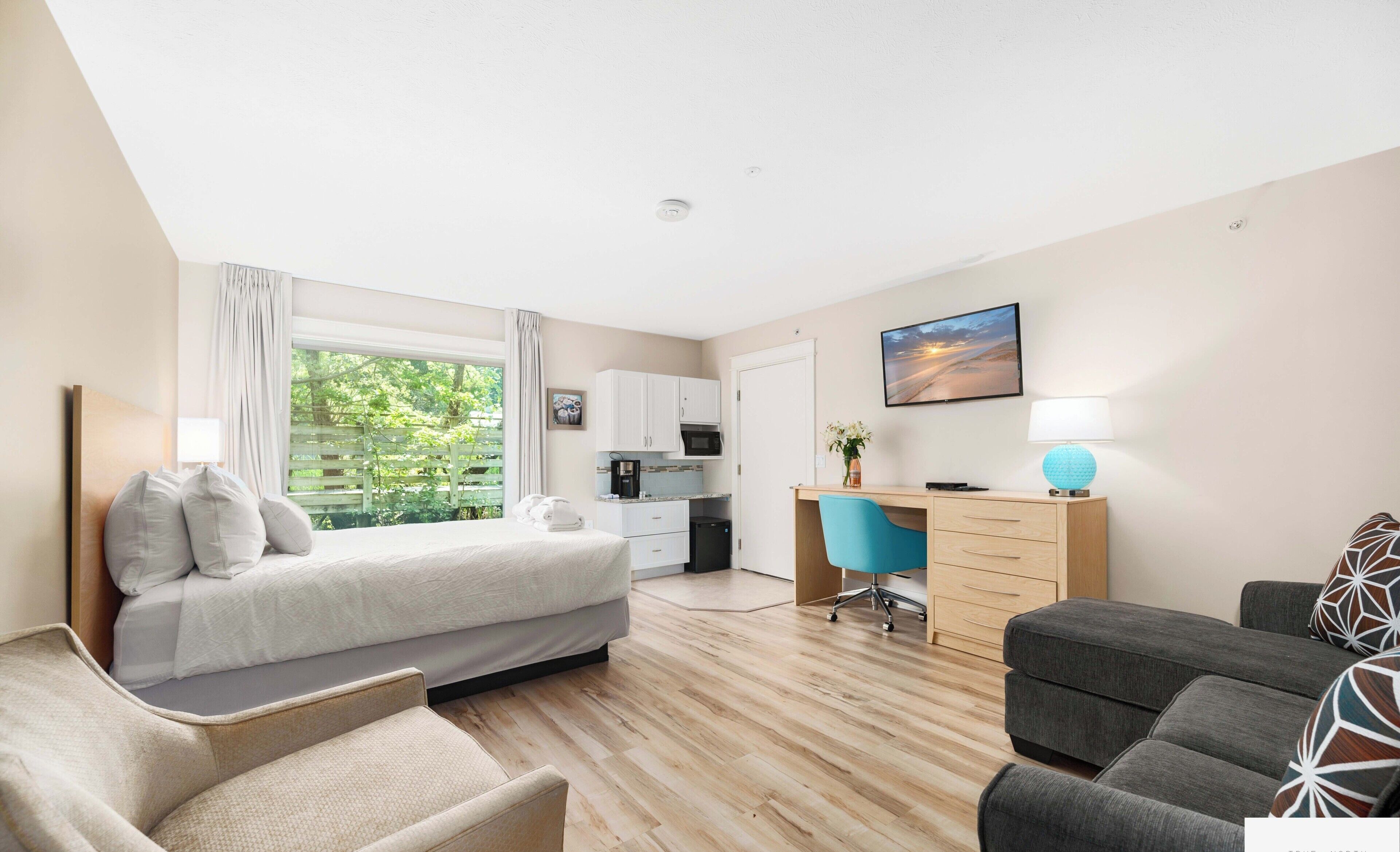 A hotel room with a bed, a desk, a chair, a sofa, a small kitchenette, and a window with curtains.