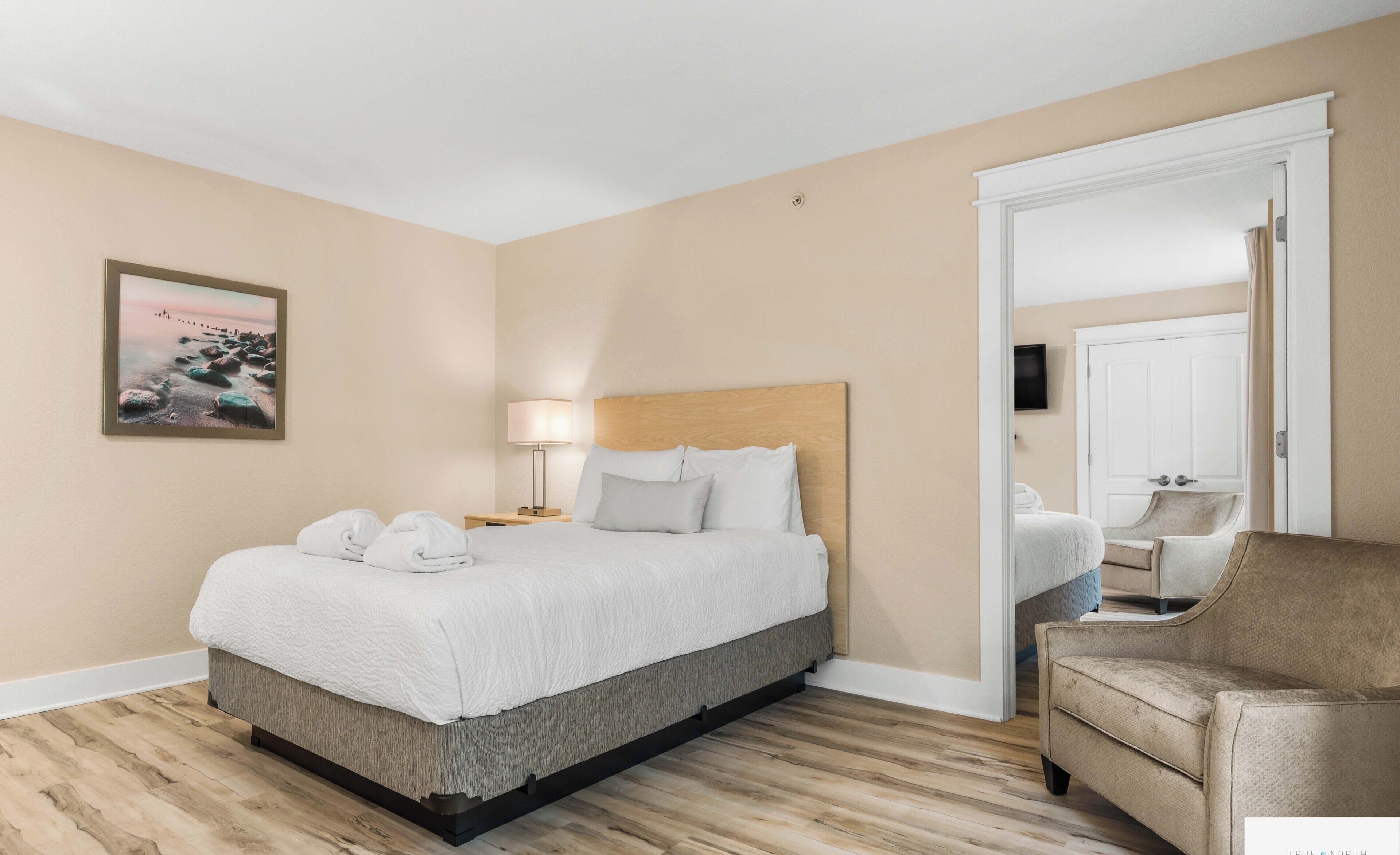 Una habitación de hotel con cama, lámpara de noche, una silla, televisor y un cuadro en la pared.