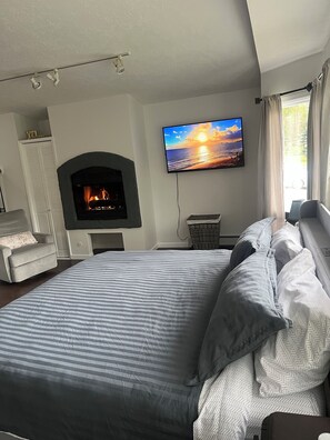 Une chambre avec une cheminée, un lit avec une literie à rayures, un téléviseur diffusant une image de coucher de soleil et une vue sur l’extérieur par une fenêtre.