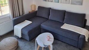 A modern living room with a grey sectional sofa, a small white table with two pink cups, a woven pouf, and a wall with framed beach and palm tree pictures. - Cottage in Zeeland Near the Beach (Koudekerke)