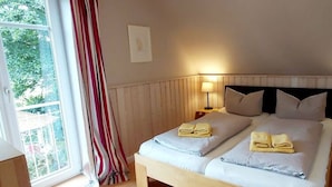 Un lit double avec deux oreillers et une table de chevet avec une lampe, une fenêtre avec des rideaux et une vue sur la verdure à l’extérieur.