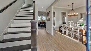 Un intérieur moderne avec un escalier, un coin repas avec des chaises blanches et un lustre, et une vue sur le salon.