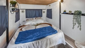 A single bed with a blue blanket, white bedding, and a wooden headboard. There are two wall-mounted shelves with plants, and a hanging plant on the right. - Cozy (sta)caravan in Pernes-les-Fontaines (Pernes-les-Fontaines)