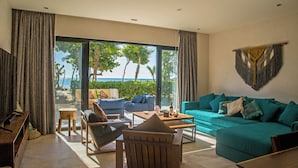 A spacious living room with a large window offering a view of the ocean and palm trees. There is a teal sofa, a wooden coffee table, and a wooden chair. - BT Homes - Tankah Beachfront (Tulum)