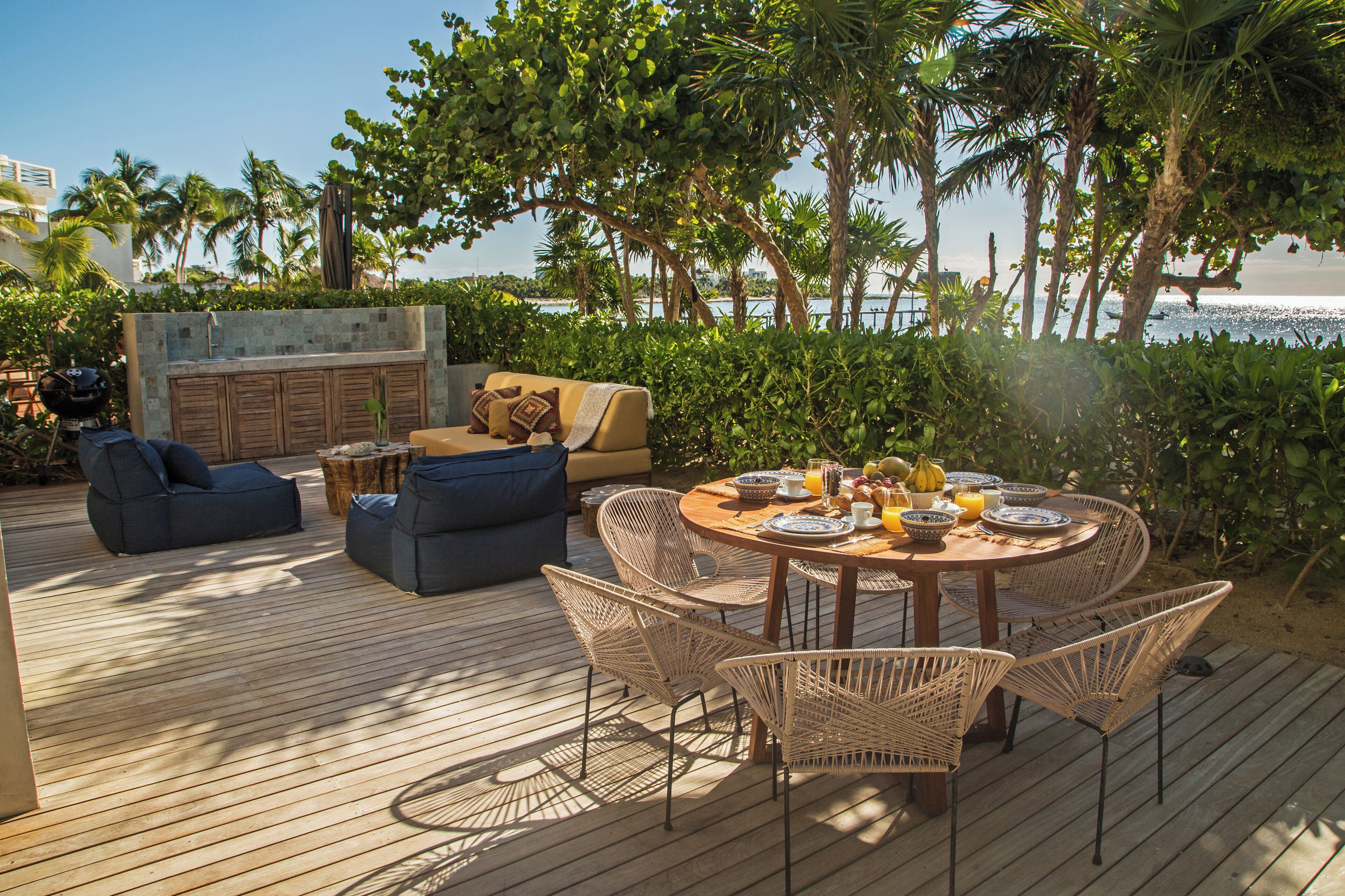 An outdoor deck with a dining table set for four, surrounded by wicker chairs, a sofa, and a grill.