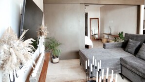 A modern living room with a grey sofa, a flat-screen TV mounted on the wall, and a potted plant. - BT Homes - Tankah Beachfront (Tulum)