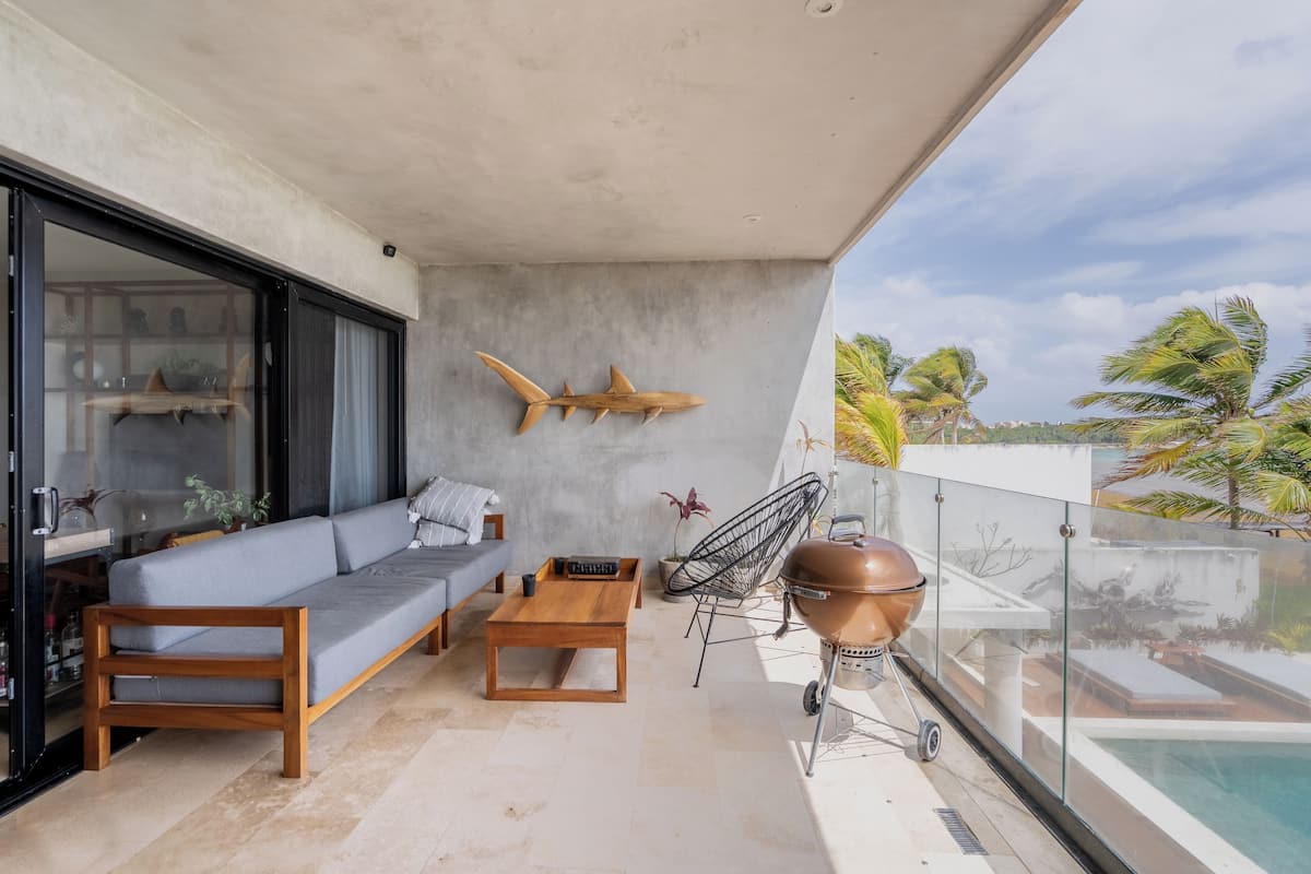 Un balcon équipé d’un canapé, d’une table basse, d’un barbecue et offrant une vue sur une piscine et des palmiers.