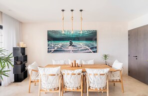 Un comedor con mesa de madera, sillas blancas acolchadas, una estantería negra y un cuadro abstracto grande en la pared.