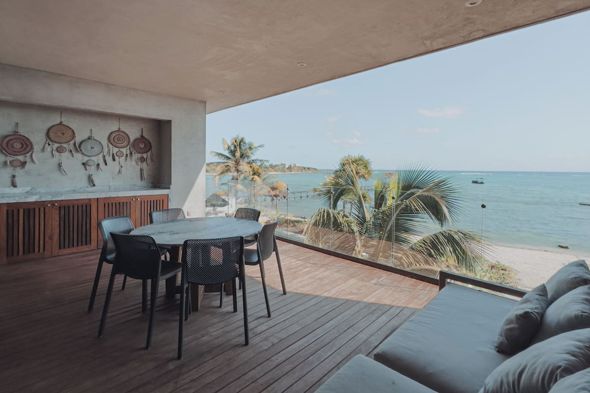 Un espace de restauration extérieur moderne avec vue sur la mer, comprenant une terrasse en bois, une table ronde avec des chaises et un mur décoré d’attrape-rêves.