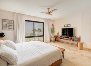 Un dormitorio con una cama grande, televisor, ventilador de techo, ventana con vista a la playa y una planta.