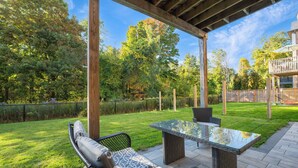 A covered patio with wicker furniture and a glass-top table, surrounded by a well-maintained lawn and trees. - Unique Industrial designer studio (Aurora)