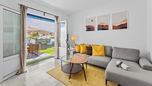 A modern living room with a grey sofa, yellow cushions, a glass coffee table, and a view of a green outdoor area through a sliding glass door.
