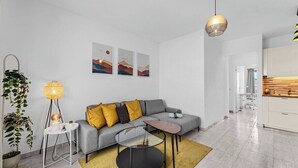 A modern living room with a grey sofa, a glass coffee table, and a yellow lamp. There are three framed pictures on the wall and a plant on the left.