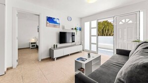 Una sala de estar moderna con un sofá gris, una mesa de centro blanca, un televisor sobre un mueble blanco y un ventanal con vistas a cactus.